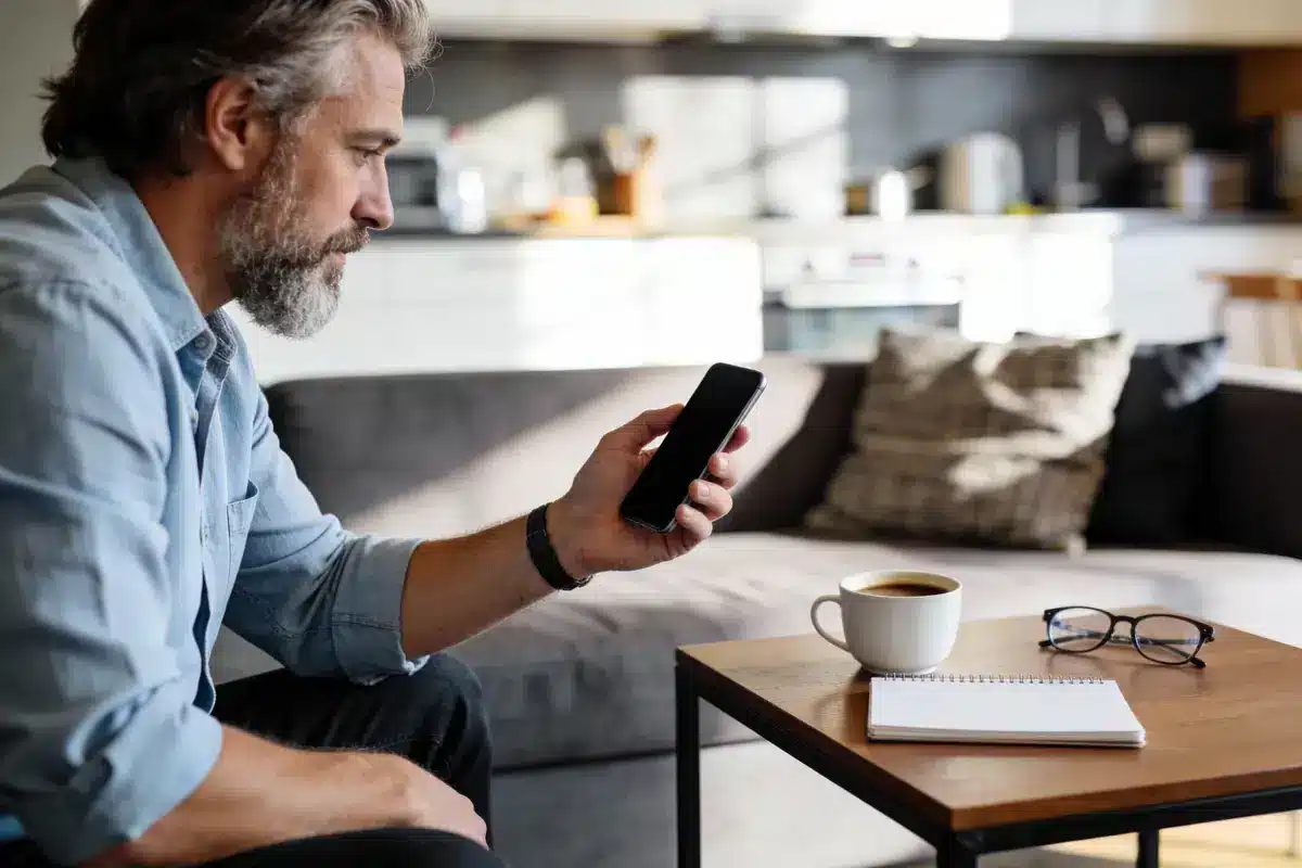 Homme regardant son smartphone, assis sur un canapé. Tasse de café et cahier sur la table.